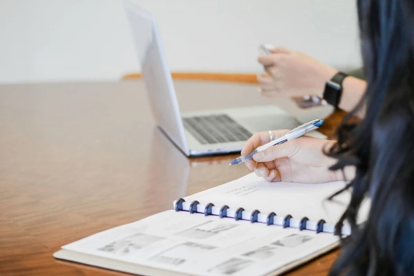 A woman seated at a table, focused on her notebook and laptop, engaged in work or study.